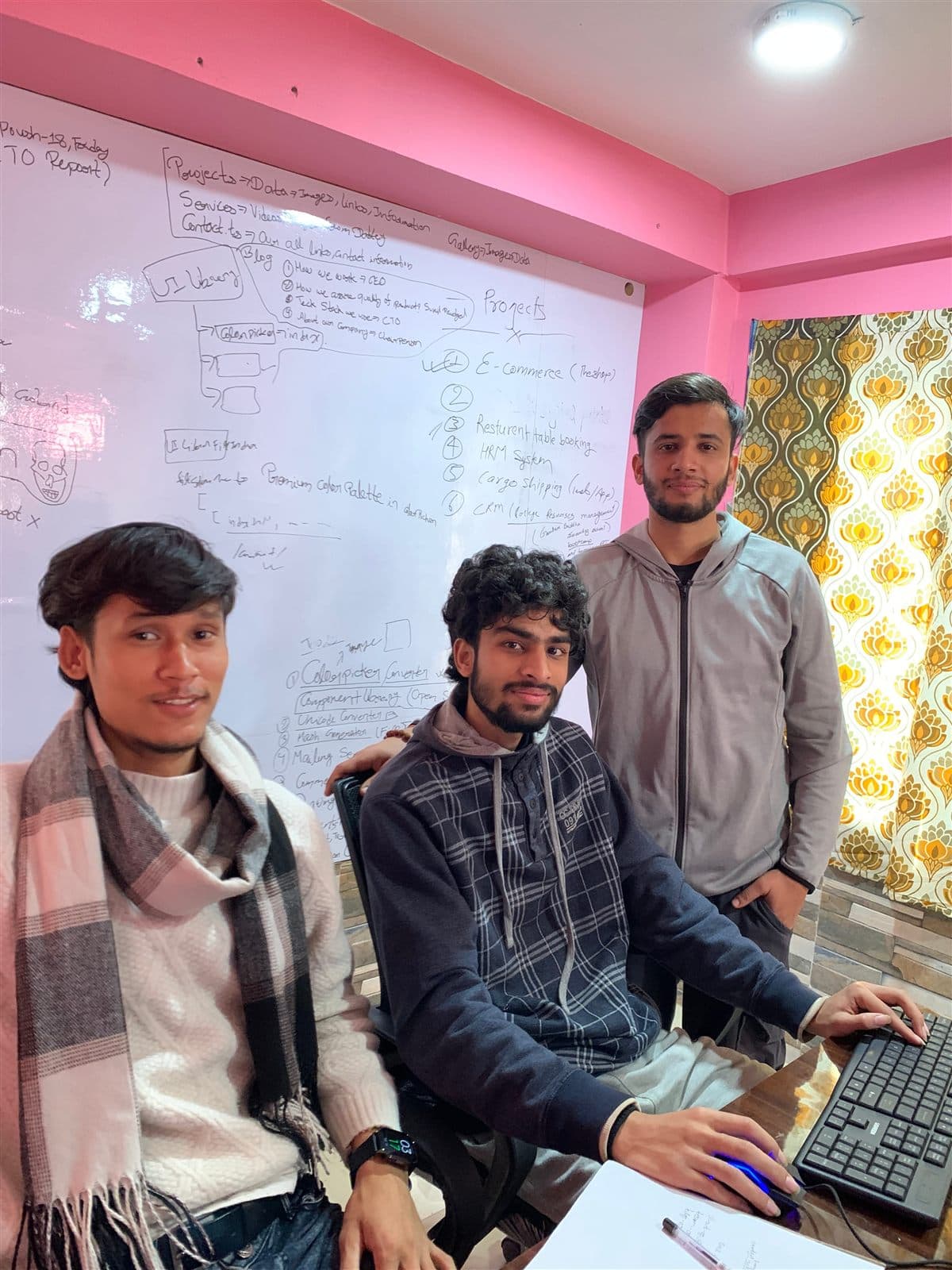 The whiteboard, mid-plan - Three of the team at the desk. On the board behind them, a numbered shortlist: e-commerce, table booking, HRM, cargo shipping, CRM.